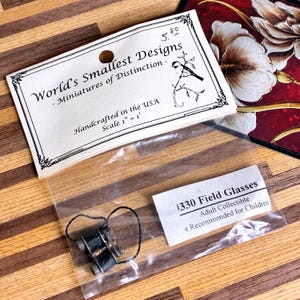 May include: Miniature field glasses, part of the "World's Smallest Designs" collection, are displayed in a clear plastic bag. The label indicates they are handcrafted in the USA, with a scale of 1" = 1'. The product is an adult collectible.