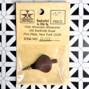 May include: A miniature bellows, handcrafted in the USA, is displayed in a clear plastic bag attached to a tan paper tag. The tag includes the text "Harmony Forge" and "Olde Mountain Miniatures". The bellows is brown with a silver nozzle.