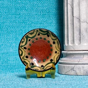 May include: A small circular ceramic dish featuring a central terracotta-colored circle surrounded by a ring of green dots and black accents. The outer rim displays a wavy green pattern on a beige background, supported by a gold-toned metal stand.