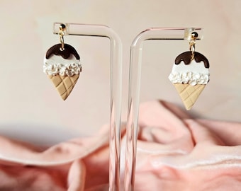 Puede incluir: Un par de pendientes originales con forma de conos de helado. Cada pendiente presenta una cobertura de chocolate marrón, helado blanco y un cono de gofre marrón claro. Los pendientes se exhiben en un soporte transparente sobre un fondo rosa.