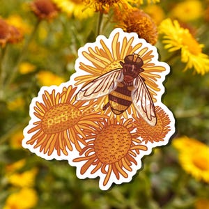 Puede incluir: Una pegatina que representa una abeja con rayas marrones y amarillas, posada sobre un grupo de flores naranjas y amarillas. La pegatina tiene un borde blanco y se encuentra sobre un fondo borroso de flores amarillas y follaje verde.