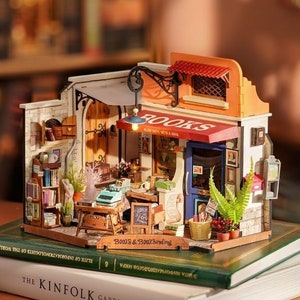 Könnte beinhalten: Ein Miniaturmodell aus Holz eines Buchladens mit einer roten Markise, auf der "BOOKS" steht, und einem Schild mit der Aufschrift "Books & Bookbinding". Das Modell steht auf zwei Büchern.