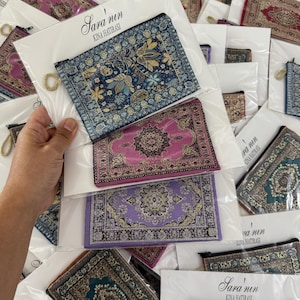 May include: Several small, rectangular fabric pouches with detailed carpet patterns in shades of blue, pink, and purple. Each pouch has a gold-coloured ring and is packaged with a white card that reads "Sara'nın Kına Hatırası."