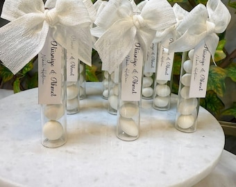 May include: Five clear glass tubes with white ribbon bows and white labels with the text "Hüsniye & Ahmet Nikah Hatırası" are filled with white candy. The tubes are arranged on a white marble surface.