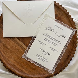 Könnte beinhalten: Eine cremefarbene Hochzeitseinladung mit weißer Spitzenborte und passendem Umschlag mit geprägten Initialen und dem Datum 19.09.2026. Der Text enthält die Namen Dilara & Selim, Veranstaltungsdetails und das Datum 19 | 09 | 2026.
