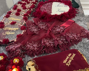 Puede incluir: Un conjunto de noche de henna en color burdeos y dorado con una cesta decorativa, una bandeja con monedas de oro, una almohada con el texto "Merve'nin Kinasi" y una bufanda con el texto "Merve'nin Kina Gecesi".