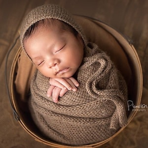 May include: A newborn baby is swaddled in a brown knitted blanket and a matching knitted hat. The baby is sleeping peacefully in a wooden bucket.
