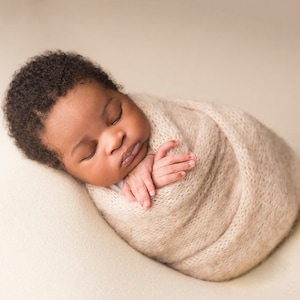 Może przedstawiać: Noworodek śpi spokojnie, owinięty w miękki, beżowy, dziergany pled. Dziecko ma ciemną skórę i ciemne włosy.