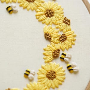 May include: Hand-stitched embroidery art showing a crescent of yellow sunflowers with brown centres and small, white and yellow bees. The embroidery is on cream-coloured fabric, stretched within a wooden hoop. A charming, handmade floral design.