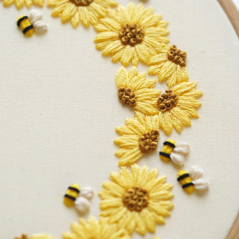 May include: Hand-stitched embroidery art showing a crescent of yellow sunflowers with brown centres and small, white and yellow bees. The embroidery is on cream-coloured fabric, stretched within a wooden hoop. A charming, handmade floral design.