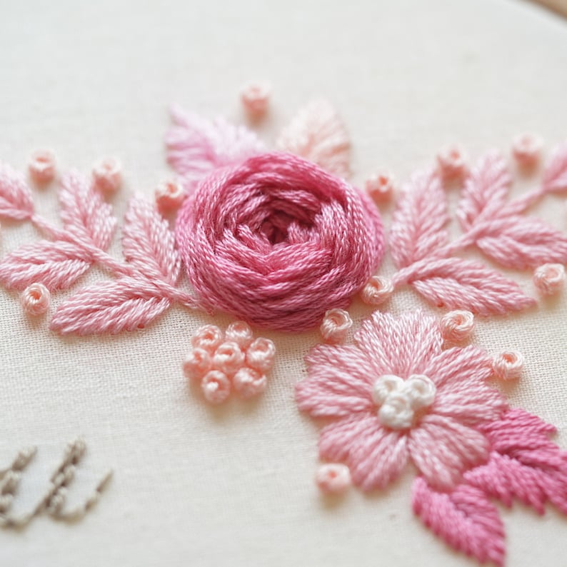 May include: Close-up of a hand-stitched embroidery featuring a central pink rose surrounded by pink leaves and smaller floral accents. The embroidery is set against a cream-colored fabric, showcasing the delicate threadwork and textures.