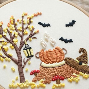 May include: An embroidery hoop featuring a brown tree with yellow leaves, a hanging lantern, three black bats, a steaming orange pumpkin, two red and white mushrooms, and a brown pointed hat on a cream-colored fabric background.