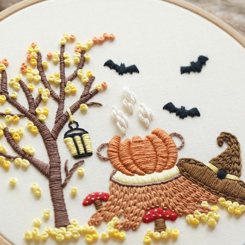 May include: An embroidery hoop featuring a brown tree with yellow leaves, a hanging lantern, three black bats, a steaming orange pumpkin, two red and white mushrooms, and a brown pointed hat on a cream-colored fabric background.