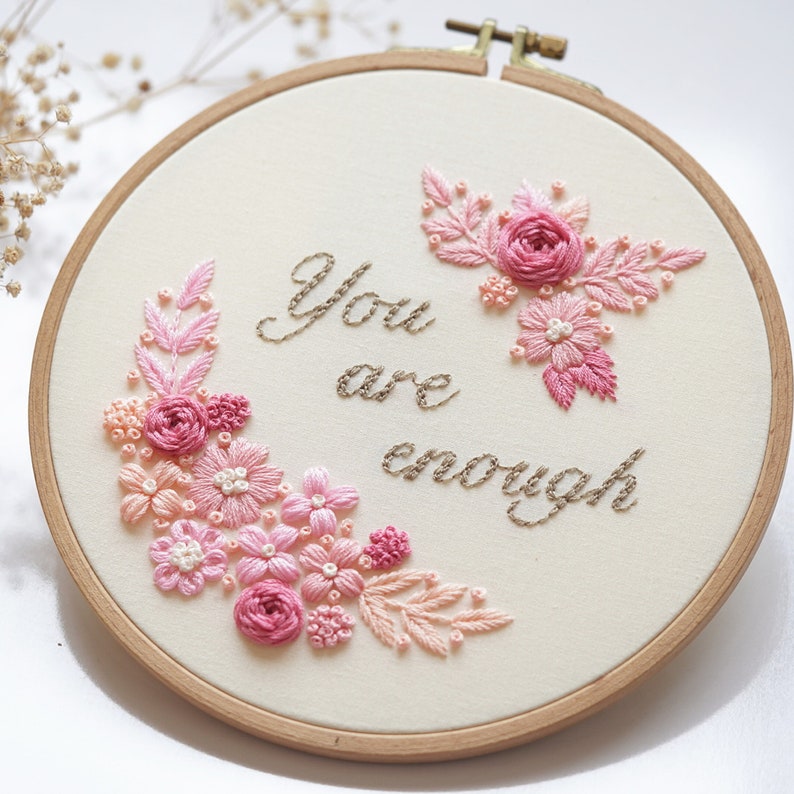 May include: A hand-stitched embroidery hoop featuring the phrase "You are enough" in brown thread. The design includes pink and peach-colored floral embroidery, with roses and other flowers, on a cream-colored fabric. The wooden hoop is visible.