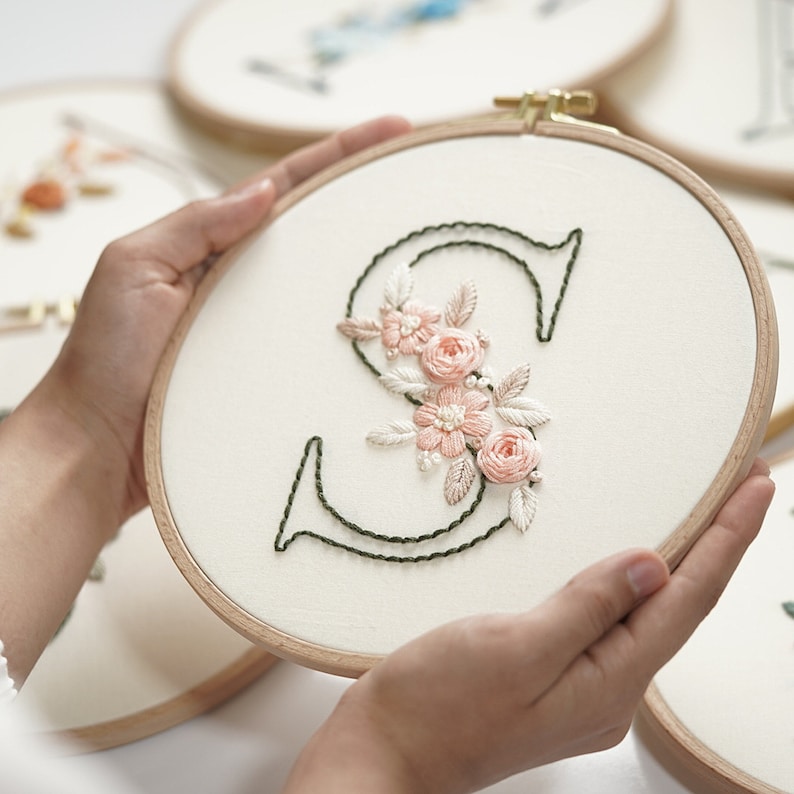 Op de afbeelding: Geborduurde hoepelkunst met de letter "S" in donkergroene draad, versierd met delicate roze en cr&egrave;mekleurige bloemenborduurwerk. Het ontwerp is geplaatst op een cr&egrave;mekleurige stof, omlijst door een houten hoepel.