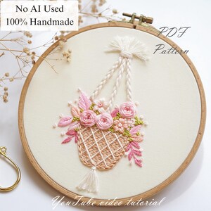 May include: Hand-stitched embroidery in a wooden hoop. The design features a hanging basket filled with pink roses and greenery. The text "No AI Used 100% Handmade" and "PDF Pattern" are visible. The words "YouTube video tutorial" are also present.