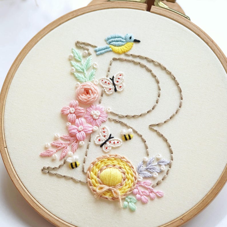 May include: Embroidered hoop art with a floral design, a blue bird, butterflies, and bees. The letter "B" is outlined with brown thread and filled with pink, yellow, and green flowers. The embroidery is set in a wooden hoop, ideal for home decor.