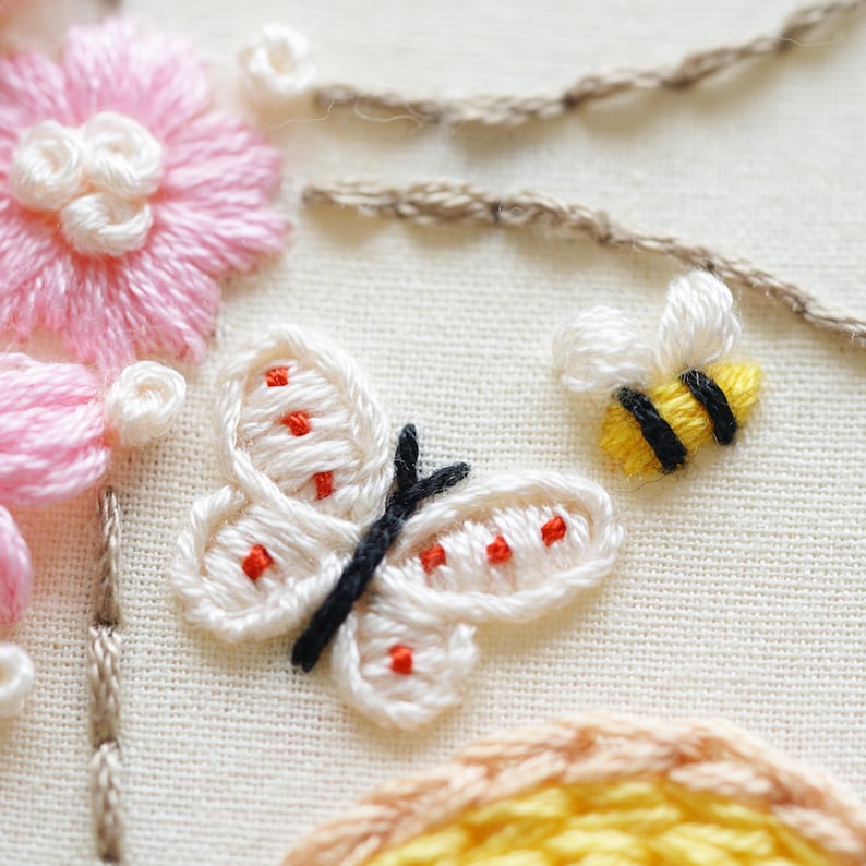 May include: Detailed embroidery showing a white butterfly with red spots and black antennae, a yellow and black bee, and pink flowers. The design is on cream fabric, with brown thread details.