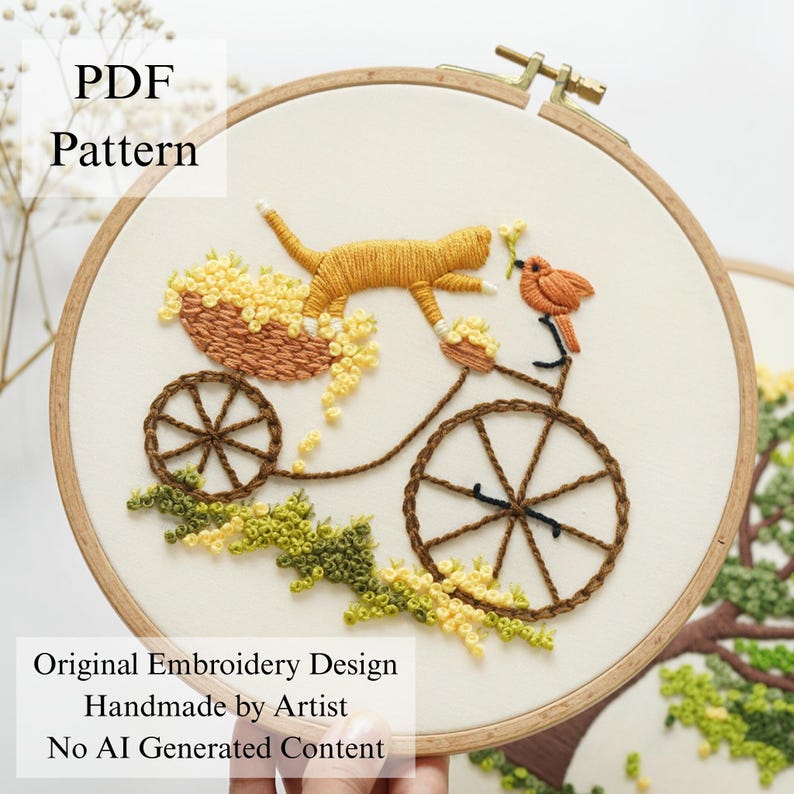 Pu&ograve; includere: Un telaio da ricamo circolare in legno contenente un pannello in tessuto con un disegno cucito a mano di un gatto arancione su una bicicletta con un cesto di fiori gialli e un piccolo uccellino arancione. Il testo PDF Pattern, Original Embroidery Design, Handmade by Artist, No AI Generated Content &egrave; visibile.