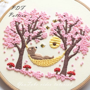 May include: Embroidered hoop art featuring a brown bear relaxing in a yellow hammock between two pink blossom trees. The trees have brown branches and are surrounded by pink flowers. There are also yellow bees and red mushrooms.