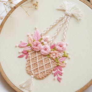 May include: Embroidered floral design in a wooden hoop. The artwork features a woven basket filled with pink roses, leaves, and small flowers. The basket is suspended by braided cords. The background is a cream-colored fabric.
