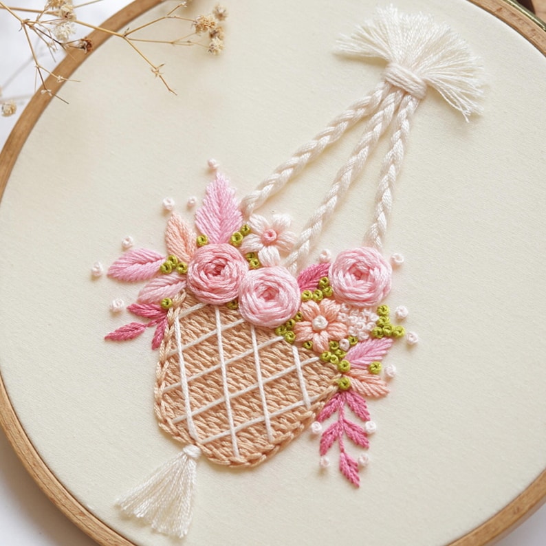 May include: Embroidered floral design in a wooden hoop. The artwork features a woven basket filled with pink roses, leaves, and small flowers. The basket is suspended by braided cords. The background is a cream-colored fabric.