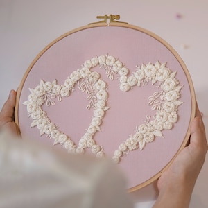 May include: A pink fabric embroidery hoop featuring two connected hearts, each outlined with white floral embroidery. The wooden hoop holds the fabric taut, creating a decorative piece. The embroidery is detailed and delicate, with a romantic theme.