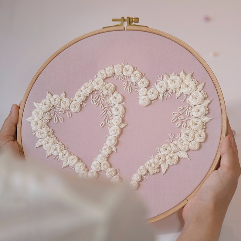 May include: A pink fabric embroidery hoop featuring two connected hearts, each outlined with white floral embroidery. The wooden hoop holds the fabric taut, creating a decorative piece. The embroidery is detailed and delicate, with a romantic theme.
