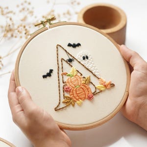 May include: A circular wooden embroidery hoop containing off-white fabric with a hand-stitched design of a brown triangular witch hat. The hat features orange pumpkins, yellow and orange leaves, a small white ghost, two black bats, and a spider.