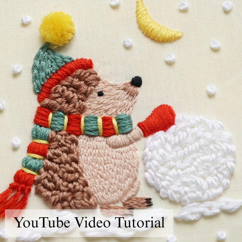 K&ouml;nnte beinhalten: Ein Stickdesign mit einem braunen Igel, der eine blaugr&uuml;ne M&uuml;tze mit gelbem Bommel und einen rot-blaugr&uuml;n-gelb gestreiften Schal tr&auml;gt. Der Igel h&auml;lt einen wei&szlig;en Schneeball unter einer gelben Mondsichel mit wei&szlig;en gestickten Schneeflocken.