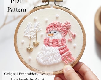 Könnte beinhalten: Ein runder hölzerner Stickrahmen mit einem weißen Stoffeinsatz, der einen handgestickten Schneemann mit einer rosa Strickmütze und einem Schal zeigt. Das Design umfasst weiße gestickte Tannenzweige, goldene Herzanhänger und kleine weiße Schneeflocken.