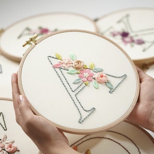 Op de afbeelding: Een borduurring met de letter "A" in grijze steken, versierd met roze, perzik en groene bloemen. De borduursel is op cr&egrave;mekleurige stof, gehouden in een houten ring. Andere ringen met bloemmotieven zijn zichtbaar op de achtergrond.