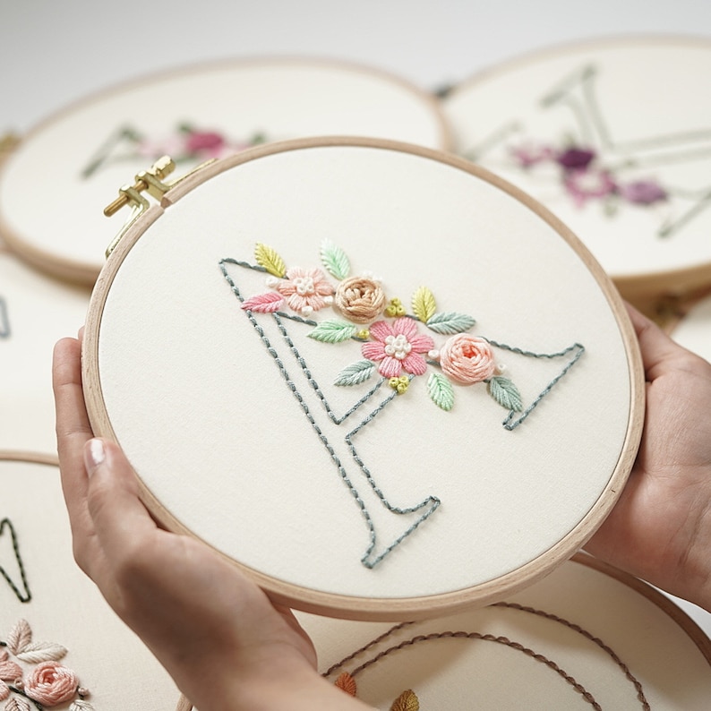 Op de afbeelding: Een borduurring met de letter "A" in grijze steken, versierd met roze, perzik en groene bloemen. De borduursel is op cr&egrave;mekleurige stof, gehouden in een houten ring. Andere ringen met bloemmotieven zijn zichtbaar op de achtergrond.