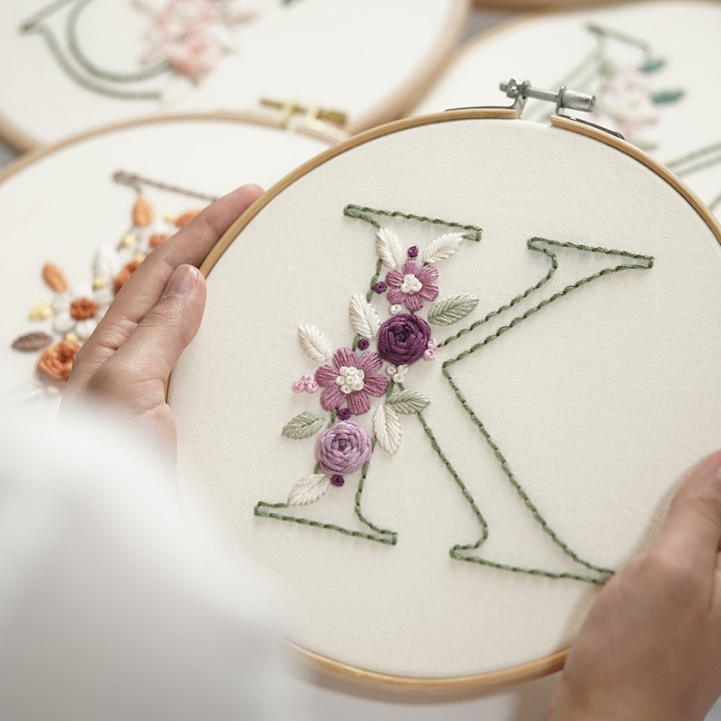 Op de afbeelding: Geborduurde letter "K" in een houten hoepel, versierd met paarse en witte bloemenborduurwerk en groene bladeren. De letter is gestikt met groen garen op een cr&egrave;mekleurige stof. Andere bloemenborduurhoepels zijn zichtbaar op de achtergrond.