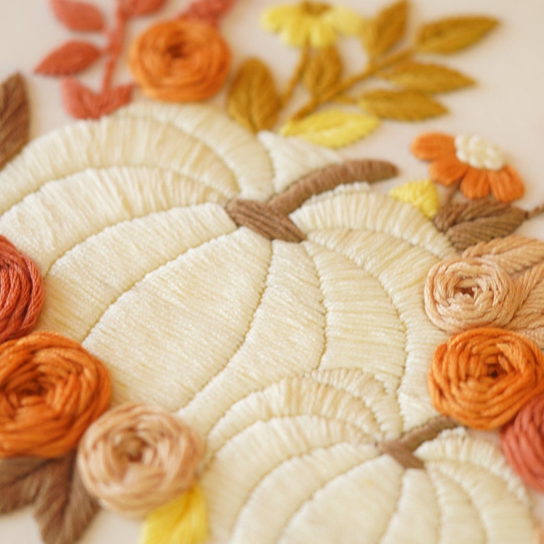 May include: An embroidery piece featuring two cream-colored pumpkins as the central focus. The pumpkins are surrounded by a decorative arrangement of embroidered flowers and leaves in shades of orange, yellow, tan, and brown thread.