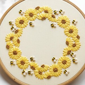 May include: Embroidered hoop art showing a wreath of yellow sunflowers and small bees. The sunflowers have detailed yellow petals and brown centres. The bees are white, black, and yellow. The background is cream-coloured fabric.