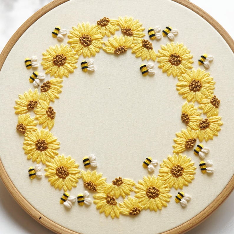May include: Embroidered hoop art showing a wreath of yellow sunflowers and small bees. The sunflowers have detailed yellow petals and brown centres. The bees are white, black, and yellow. The background is cream-coloured fabric.