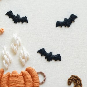 May include: An embroidery piece on off-white fabric featuring three black stitched bats in flight. Below the bats, there is an orange pumpkin with white stitched steam rising from it and a small brown curved shape on the lower right side.