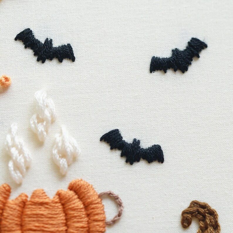 May include: An embroidery piece on off-white fabric featuring three black stitched bats in flight. Below the bats, there is an orange pumpkin with white stitched steam rising from it and a small brown curved shape on the lower right side.