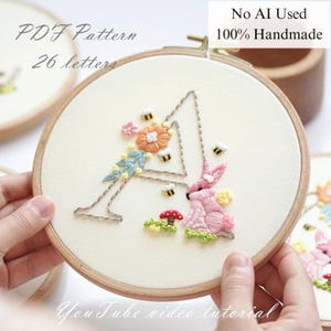 Könnte beinhalten: Gestickter Buchstabe "A" in einem Holzreifen. Das Design zeigt ein rosa Kaninchen, Blumen, Bienen und einen Pilz. Der Text "PDF Pattern 26 letters" und "No AI Used 100% Handmade" sind sichtbar. Die Worte "You Tube video tutorial" sind ebenfalls sichtbar.