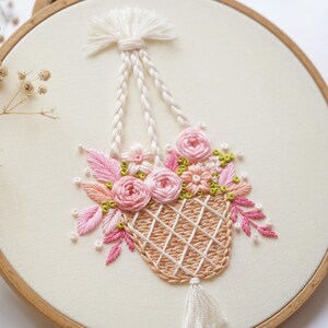 May include: Embroidered floral design in a wooden hoop. The artwork features a woven basket filled with pink roses, leaves, and small flowers. The basket is suspended by white rope. The background is a cream-colored fabric.