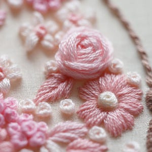 May include: Detailed embroidery showing pink and white floral designs. A central rose is surrounded by smaller flowers and leaves, all carefully stitched with thread on a light-coloured fabric. The soft colour palette creates a gentle, feminine aesthetic.