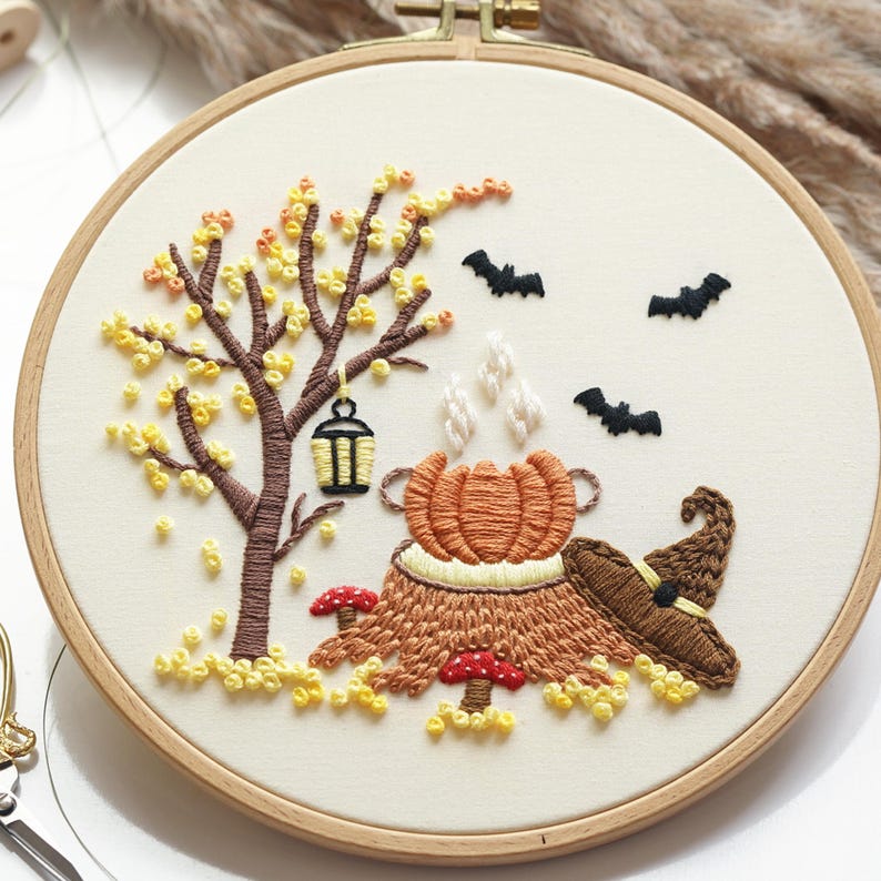 May include: A circular wooden embroidery hoop containing a fabric panel with a hand-stitched autumn scene. The design features a brown tree with yellow leaves, a hanging lantern, a steaming orange pumpkin, red mushrooms, and black bats.