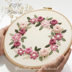 Puede incluir: Un aro de bordado blanco con un diseño de corona floral rosa y verde. El texto "PDF Pattern" y "YouTube video tutorial" es visible en el aro.