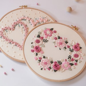 May include: Two embroidery hoops with floral designs. One features a heart shape made of pink roses, the other a wreath of pink and white flowers. Both are on cream-colored fabric, set in wooden hoops.