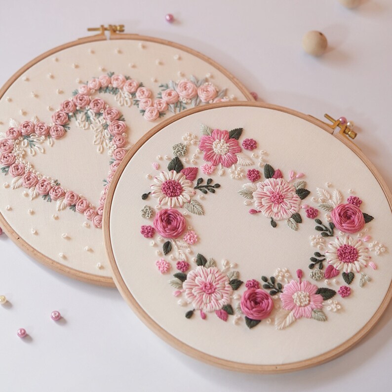 May include: Two embroidery hoops with floral designs. One features a heart shape made of pink roses, the other a wreath of pink and white flowers. Both are on cream-colored fabric, set in wooden hoops.