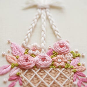 May include: Close-up of an embroidered floral basket. The basket is filled with pink roses, peach-colored flowers, and green accents. The basket has a woven design and is suspended by white braided handles. The background is a soft, off-white color.