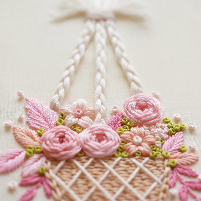 May include: Close-up of an embroidered floral basket. The basket is filled with pink roses, peach-colored flowers, and green accents. The basket has a woven design and is suspended by white braided handles. The background is a soft, off-white color.