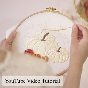 May include: A round wooden embroidery hoop holding white fabric with an unfinished needlepoint design of two pumpkins and floral outlines. A gold needle with yellow thread is positioned above the pumpkins, which are stitched in cream and brown.