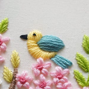 May include: A close-up of an embroidered design featuring a yellow bird with a blue wing and tail, a black beak, and a black eye. Pink flowers and green leaves surround the bird. The embroidery is on a white fabric background.
